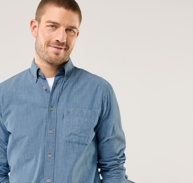 Ein Mann trägt ein hellblaues Jeanshemd über einem weißen T-Shirt und lächelt leicht. Der Stil wirkt locker und modern.