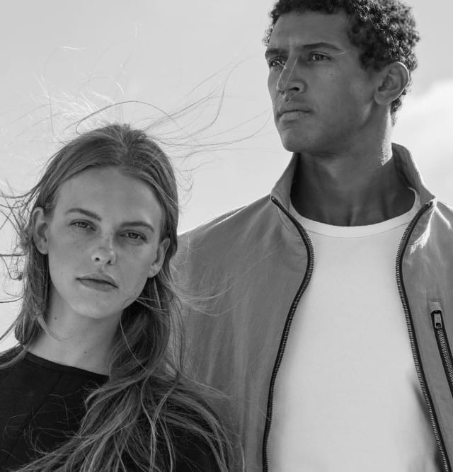 Zwei Personen stehen nebeneinander vor einem hellen Himmel. Eine Frau trägt ein schlichtes, dunkles Oberteil, ein Mann trägt eine helle Jacke über einem weißen Shirt. Der Stil ist modern, minimalistischen Mode-Stil mit klarem, zeitlosem Ausdruck.