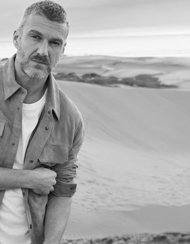 Ein Mann steht in einer Dünenlandschaft und trägt ein helles T-Shirt unter einer offenen Hemdjacke mit Brusttaschen.