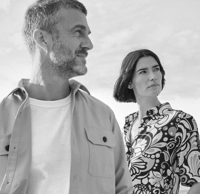 Zwei Personen stehen nebeneinander in den Dünen. Eine Frau trägt eine gemusterte Blusen, ein Mann trägt eine Hemdjacke über einem weißen Shirt. Der Stil ist modern, minimalistischen Mode-Stil mit klarem, zeitlosem Ausdruck.