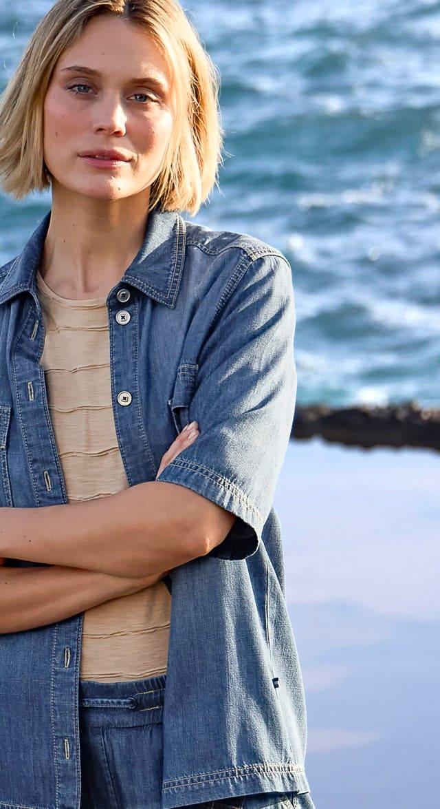 Eine Frau trägt ein blaues Jeanshemd über einem beigefarbenen Oberteil und steht mit verschränkten Armen am Meer.