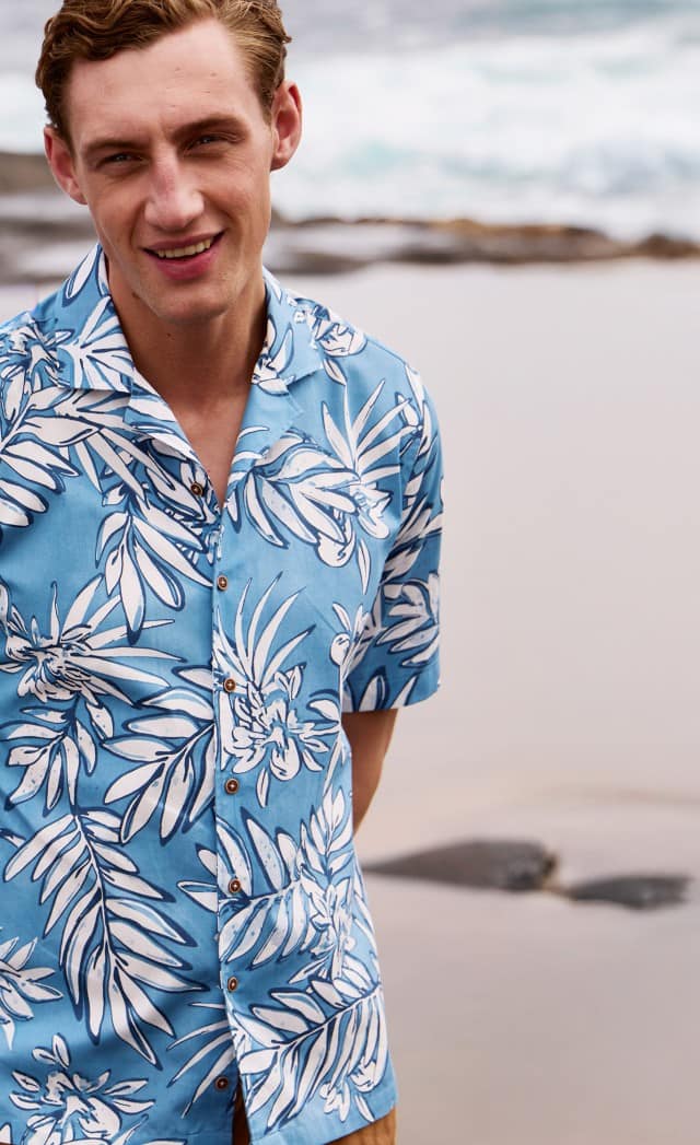 Ein Mann trägt ein blau-weiß geblümtes Kurzarmhemd und steht an einem felsigen Strand.