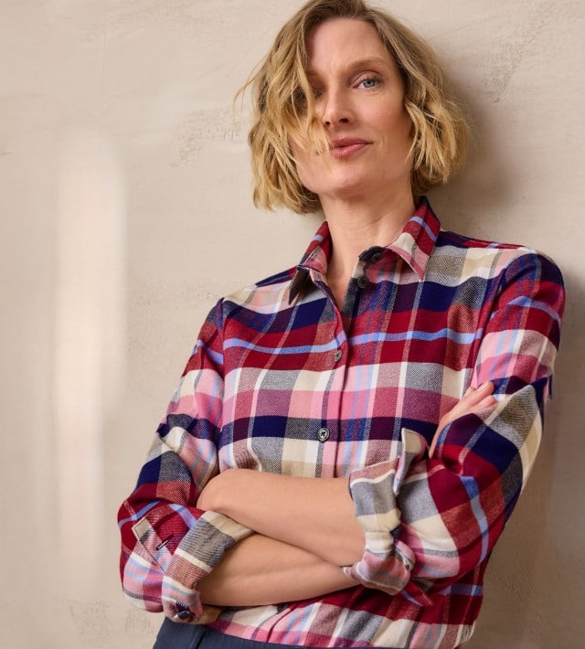 Eine Frau steht mit verschränkten Armen an einer Wand und trägt eine karierte Thermo-Flanell-Hemdbluse.