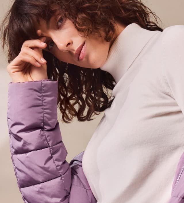 Eine Frau trägt einen weißen Rollkragenpullover mit passender heller Hose. Der Look ist elegant, minimalistisch und mit einer lila Steppjacke modern akzentuiert.
