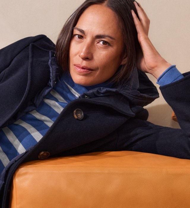 Frau in einem gestreiften blau-grauen Pullover, Jeans und einem dunkelblauen Mantel, sitzt lässig auf einer Couch und schaut in die Kamera.