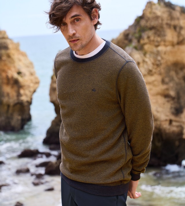 Ein Mann steht vor einem Meer mit Felsen, trägt einen Strukturpullover in Braun- und Blautönen über einem weißen T-Shirt und Jeans und schaut in die Kamera.
