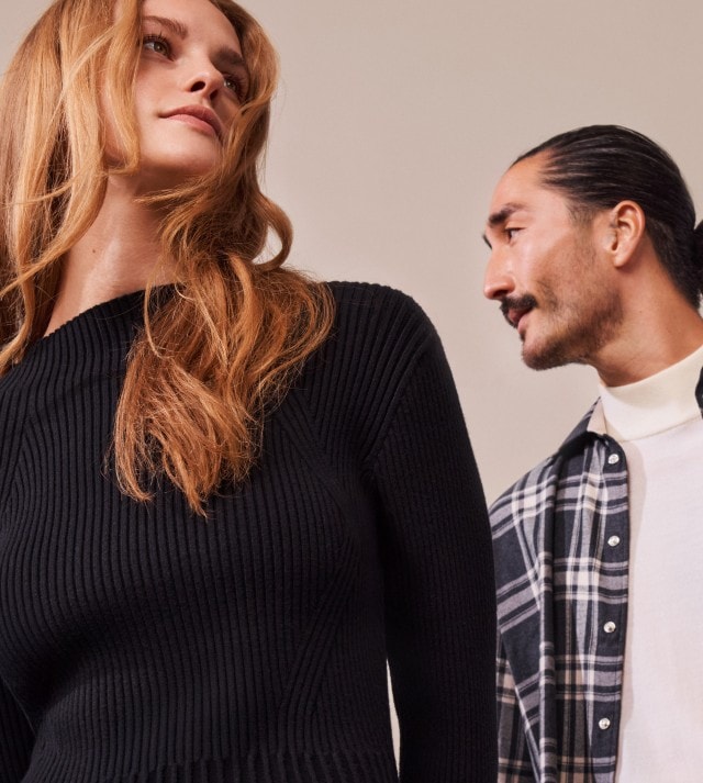 Eine Frau in einem schwarzen Strickpullover und ein Mann in weißem Rollkragen und offenem schwarz-weiß karierten Hemd stehen vor einem neutralen Hintergrund.