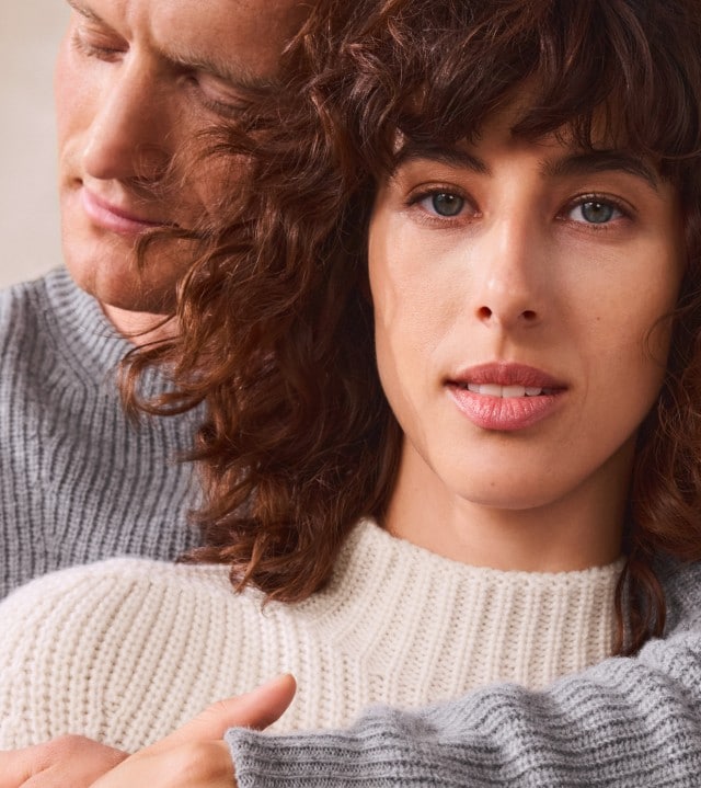 Eine Frau in einem weißen Rollkragenpullover wird von einem Mann in einem grauen Strickpullover umarmt und schaut in die Kamera.