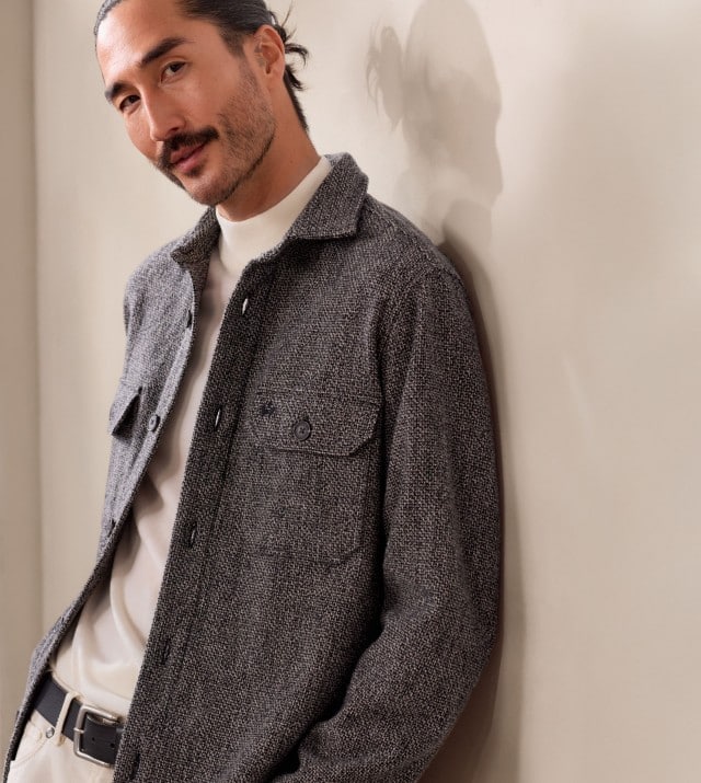 Ein Mann trägt ein helles Oberteil mit hohem Kragen und darüber eine graue, grob strukturierte Hemdjacke mit Knöpfen und einer Brusttasche.