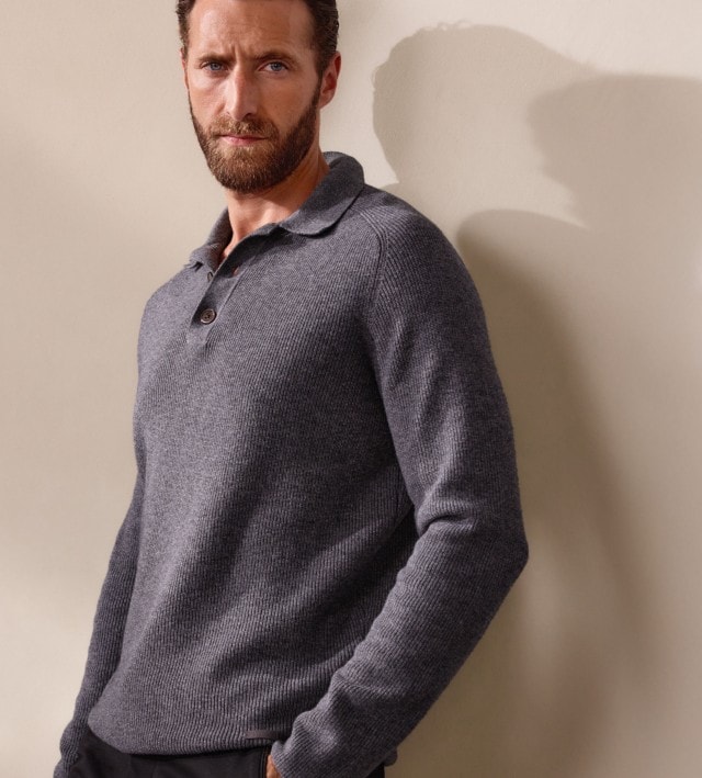 Mann in einem grauen Strickpullover lehnt an einer hellen Wand und schaut in die Kamera.
