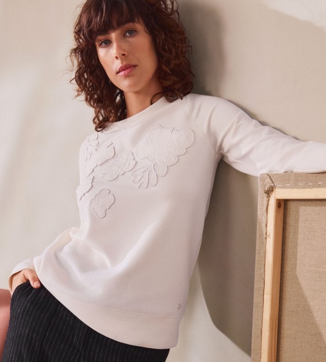 Frau in einem weißen bestickten Pullover und gestreifter dunkler Hose lehnt lässig an einer weißen Wand.