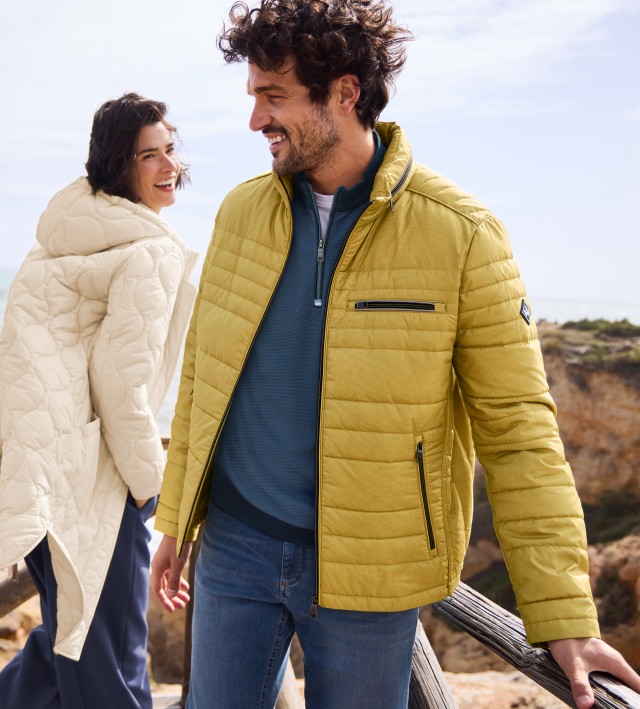 Im Fokus ist ein Mann, der mit einer Frau auf Felsen an einem Meer steht, eine gelbe Leichtjacke über einem blauen Troyer trägt und lachend zur Seite schaut.