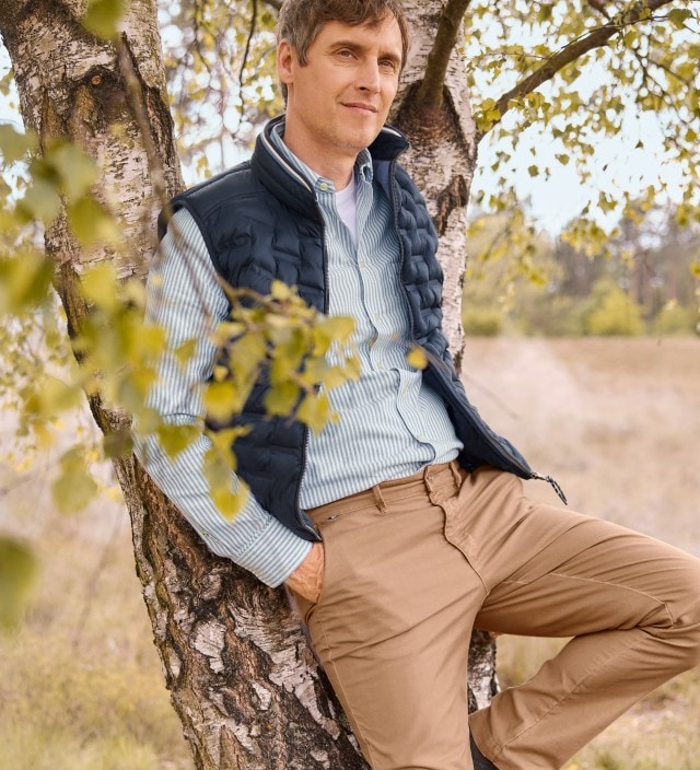 Ein Mann, an einem Baum angelehnt, trägt ein gestreiftes Hemd, darüber eine blaue Steppweste und dazu eine Hose in Beige.