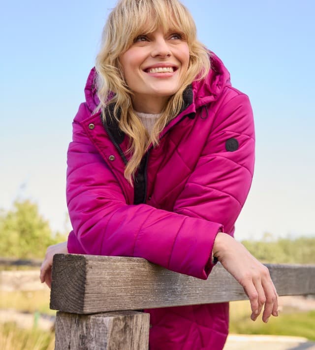 Ein Frau, an einem Zaun gelehnt, trägt eine Aquastop Thermojacke in der Farbe Waldbeere, darunter einen weißen Strickpullover. 