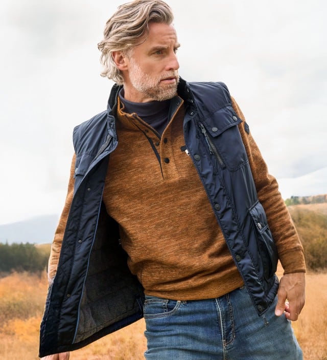 Ein Mann trägt einen rostbraunen Pullover mit Knopfleiste und eine dunkelblaue Steppweste zu Jeans; der Look wirkt lässig und herbstlich-sportlich.