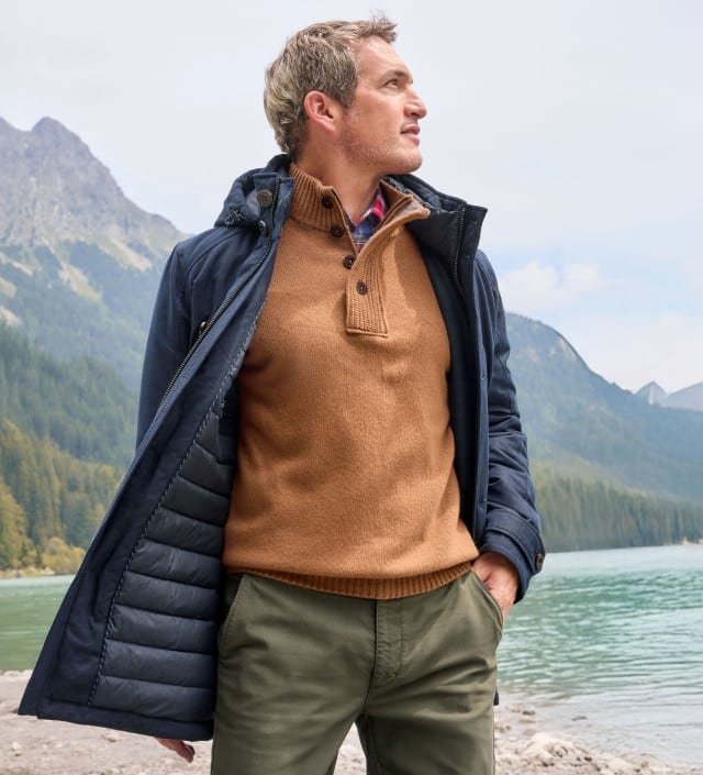 Ein Mann steht vor einem Gebirge, schaut zur Seite und trägt den Ultraskin Leichtparka in Marine, darunter einen Premium Lammwoll-Troyer in Camel und dazu eine Hose in Oliv.