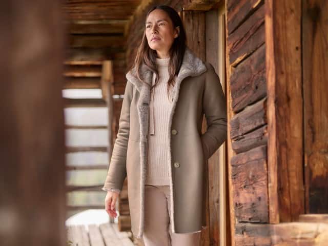 Eine Frau steht vor einer Hütte und trägt einen Rollkragen-Pullover, darüber den Premium Lammfell Wendemantel in Taupe. Eine Frau steht vor einer Hütte und trägt einen Rollkragen-Pullover, darüber den Premium Lammfell Wendemantel in Taupe.