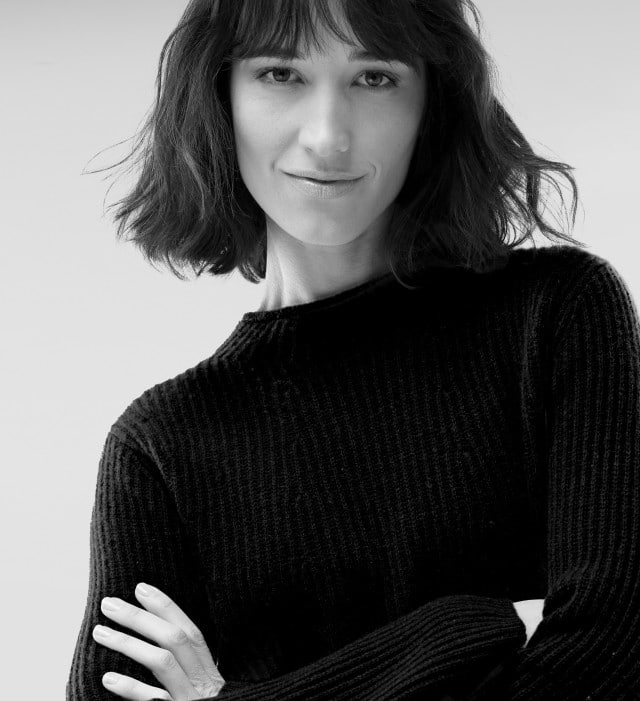 Eine Frau trägt einen dunklen Strickpullover mit Stehkragen und verschränkt die Arme.