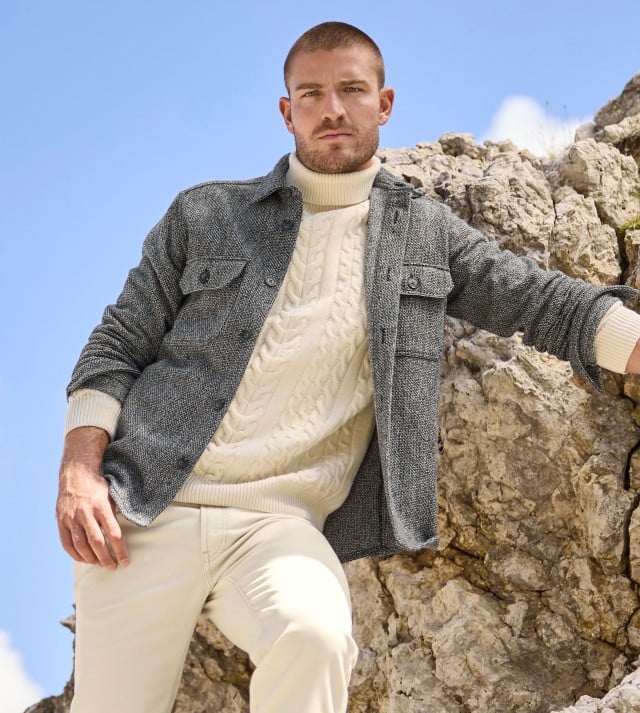 Ein Mann steht auf einem Felsen und trägt eine Hemd Jacke in Schwarz, darunter einen Rollkragen Pullover mit Zopfmuster in Offwhite und dazu eine helle Hose.