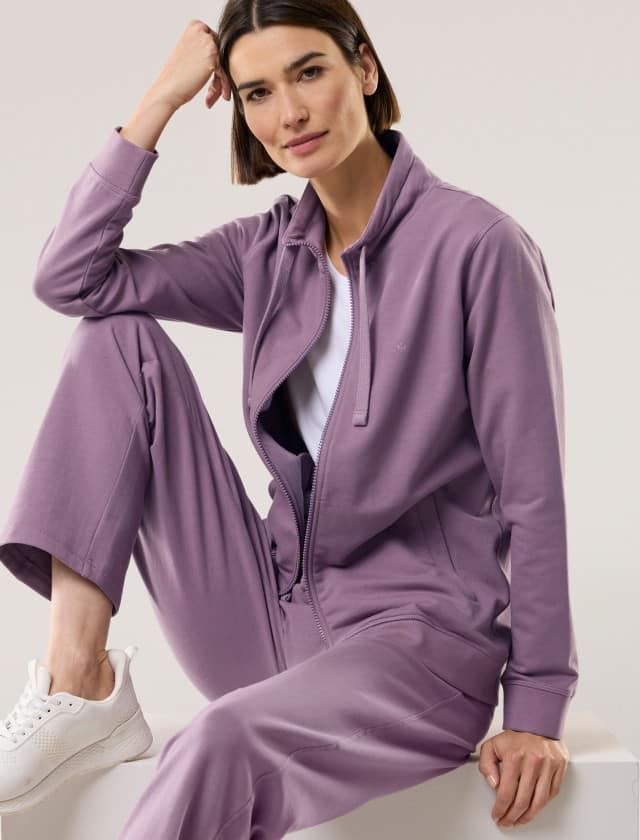 Person trägt ein hellviolettes Freizeit-Set aus Sweatshirtjacke und passender Hose sowie ein weißes T-Shirt und weiße Sneaker in sportlichem Stil.