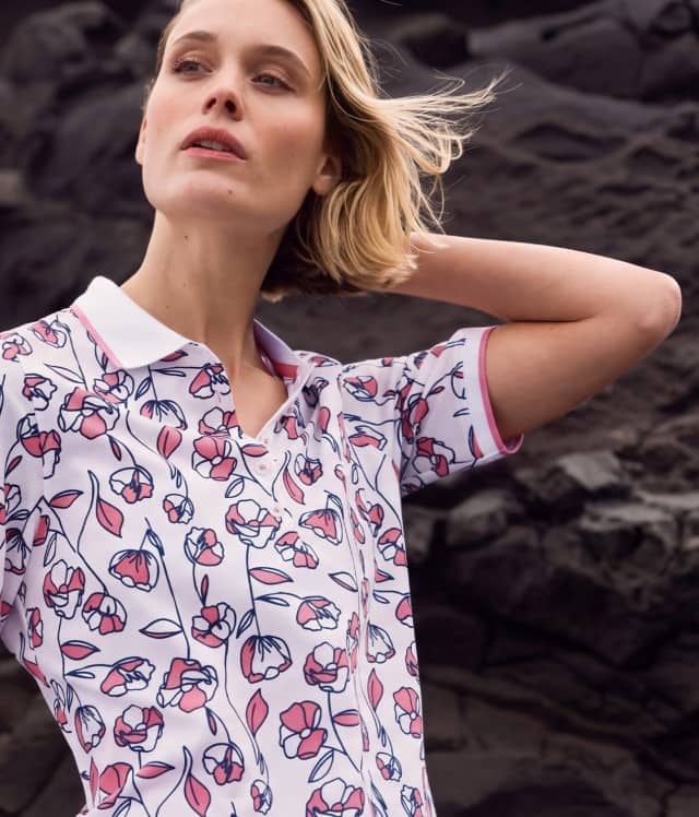 Frau mit blondem Haar in floral gemustertem Poloshirt vor einer dunklen Felslandschaft.