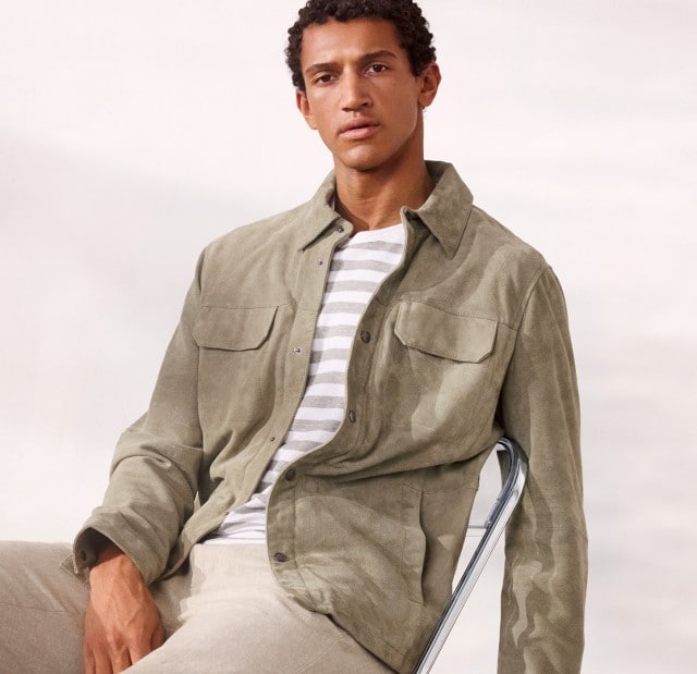 Ein Mann sitzt lässig auf einem Stuhl und trägt eine Velourslederhemdjacke in der Farbe Salbei, darunter ein gestreiftes Shirt und dazu eine helle Hose.