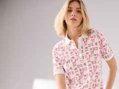 Frau mit blondem Haar trägt ein weißes Polo-Shirt mit rosa Blumenmuster vor grauem Hintergrund.
