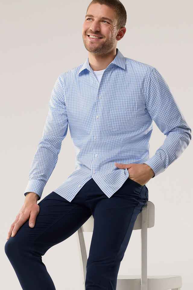 Mann in kariertem Hemd und dunkler Hose sitzt auf einem Hocker und lächelt.