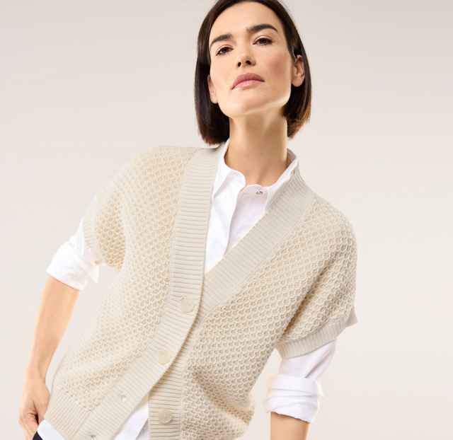 Eine Frau trägt eine Strickweste mit Waffelstruktur in Offwhite, darunter eine weiße Bluse.