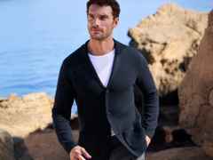 Mann in dunkelblauem Cardigan und weißem T-Shirt steht vor felsiger Küste mit Blick aufs Meer im Hintergrund.