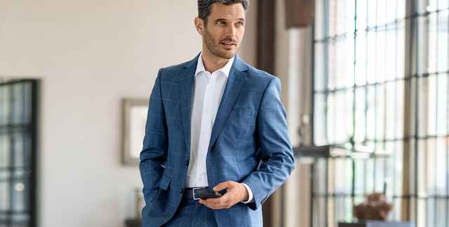 Mann steht in einem Loft und trägt einen blau karierten Anzug und hält ein Handy in der Hand.