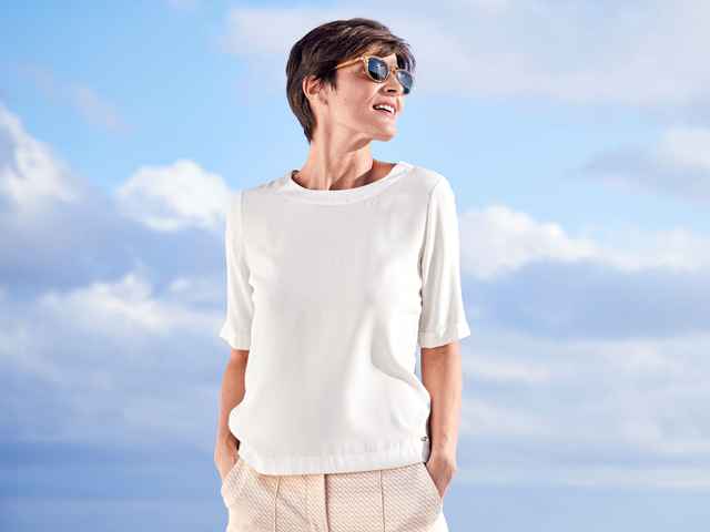 Frau mit Sonnebrille und kurzen Haaren trägt eine weiße Shirtbluse, im Hintergrund sind Wolken.
