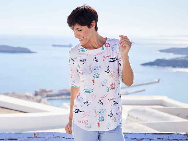 Frau steht vor dem Meer und trägt eine Kurzarm-Bluse mit Muster.