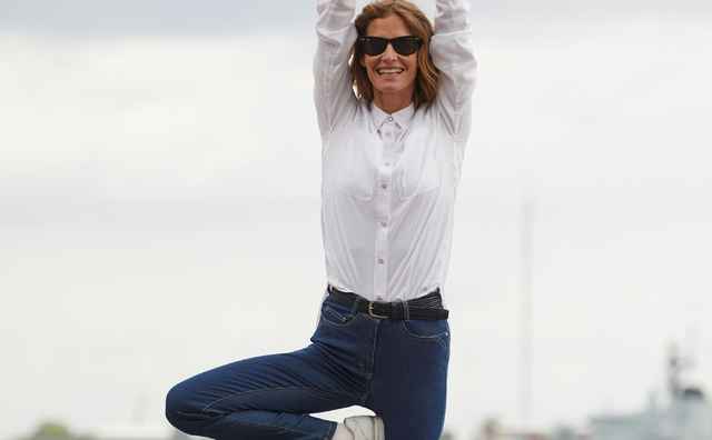 Frau im weißen Hemd und Jeans steht im Baum-Yoga-Pose am Wasser, trägt Sonnenbrille, Hintergrund verschwommene Hafenansicht.