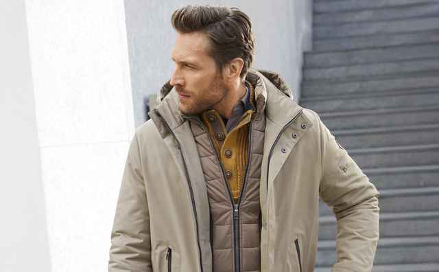 Mann in beigefarbener Winterjacke mit Kapuze, darüber braune Steppjacke; er steht draußen vor einer Treppe und schaut nach links.
