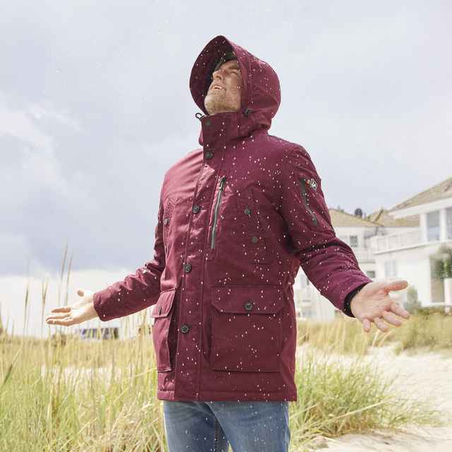 Ein Mann steht in einem weinroten Parka im Regen mit erhobenen Armen auf einem strandnahen Weg. Im Hintergrund sind Häuser und Dünen zu sehen.