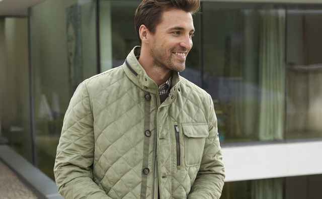 Mann in olivgrüner Steppjacke steht lächelnd auf einer Terrasse vor einem modernen Gebäude.