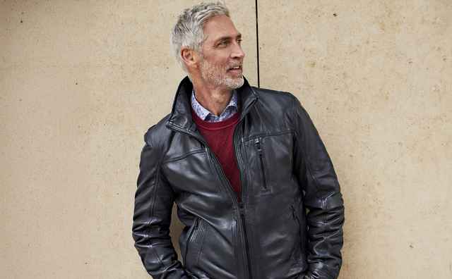 Mann mit grauem Haar trägt eine schwarze Lederjacke und einen roten Pullover, lehnt lässig an einer beigen Wand und blickt zur Seite.