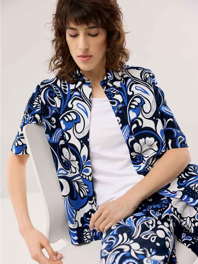 Eine Frau trägt ein blau‑weiß gemustertes Set aus Hemd und Hose über einem weißen Oberteil, was einen modernen, lebendigen und zugleich entspannten Eindruck vermittelt.