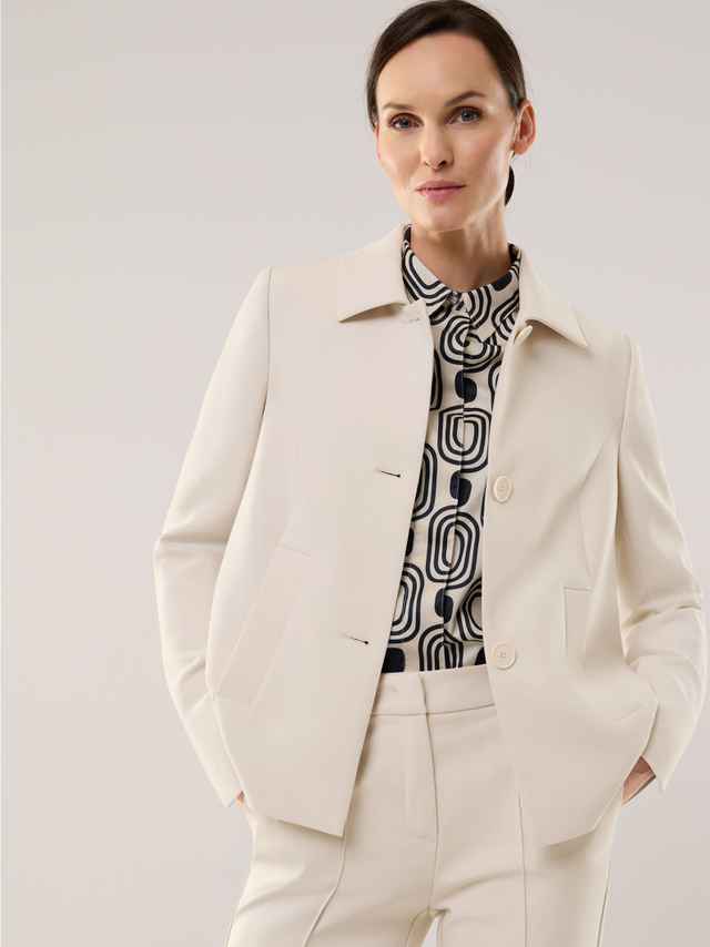 Eine Frau trägt einen eleganten cremefarbenen Hosenanzug mit einer grafisch gemusterten Bluse, das Outfit wirkt ruhig, modern und stilvoll.