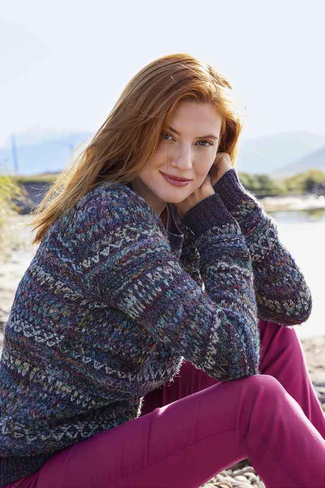 Eine Frau sitzt in einem Alpaka-Pullover in der Sonne am Strand.