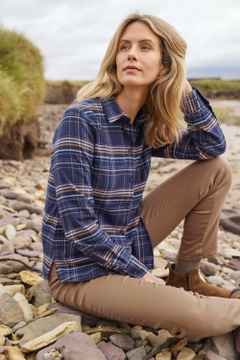 Eine Frau sitzt auf Steinen am Ufer und trägt eine karierte Flanellbluse, dazu eine Hose in Beige.