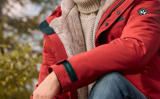 Mann mit grauen Haaren trägt rote Outdoor-Jacke und sitzt in der Natur, blickt nachdenklich in die Ferne.