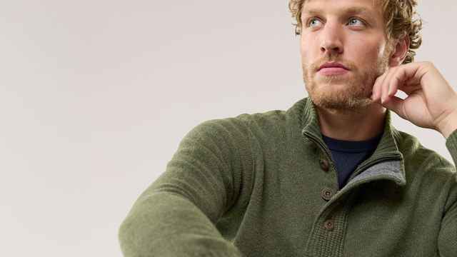Ein Mann trägt einen olivgrünen Strickpullover mit Knopfleiste und hohem Kragen. Der Stil wirkt gemütlich, natürlich und sportlich-elegant.