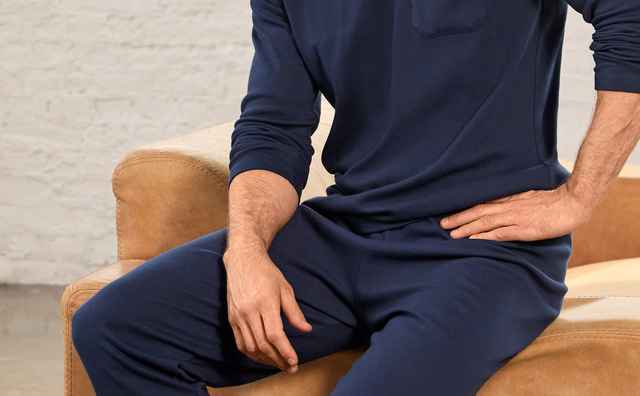 Ein Mann sitzt entspannt auf einem Sofa und trägt einen dunkelblauen, langen Schlafanzug mit V‑Ausschnitt und Brusttasche. Der Look wirkt gemütlich, schlicht und angenehm relaxed.