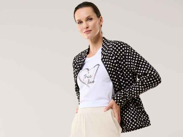 Einer Frau trägt eine gepunktete Sweatjacke, mit weißen T-Shirt und heller Hose.