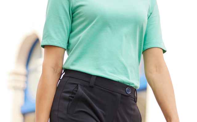 Frau in mintgrünem Shirt und schwarzer Hose spaziert mit Handtasche in einer sonnigen Straße, lächelnd und selbstbewusst.