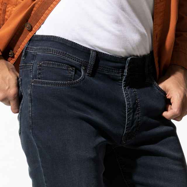 Person trägt dunkelblaue Jeans, weißes T-Shirt und orange Jacke, mit den Händen in den Hosentaschen.