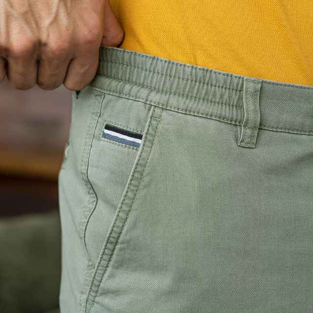 Hand zieht an einem grünen Hosenbund mit gestreiftem Etikett, gelbes Shirt im Hintergrund.