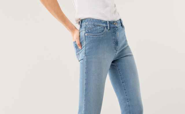 Frau in weißem T-Shirt und blauer Jeans macht einen Schritt nach vorn. Sie trägt weiße Turnschuhe und hat blondes Haar. Hintergrund ist schlicht.